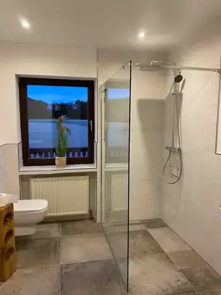 Helles Badezimmer mit großzügiger begehbarer Glasdusche, weißen Wandfliesen und Abendlicht durch das Fenster.