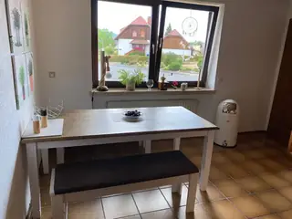Lichtdurchfluteter Essplatz mit Holztisch, gepolsterter Bank und Ausblick auf Dächer und Hügel durch das große Fenster.